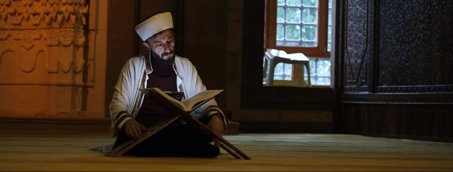 Fotografi av en mann med skjegg som sitter på gulvet og leser i en tykk bok. Boka ligger oppslått på et lite stativ foran ham. Han har på seg en hvit jakke med gullpynt på og et hvitt hodeplagg. Bak ham ser man veggen og vinduet til moskeen han sitter i.