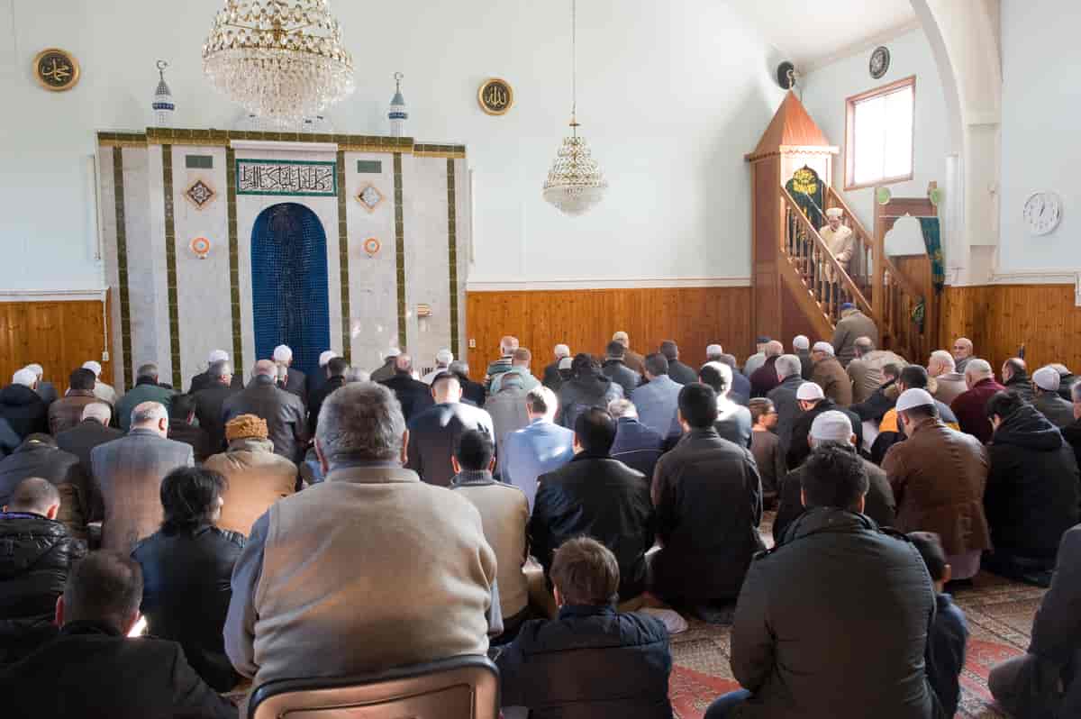 Fotografi av en gruppe menn som ber i en moské. De sitter på gulvet med ryggen til kamera. Foran dem står et slags stort, bredt skap. Foran dem til høyre står en mann i en slags liten trapp. Han er vendt mot mennene som ber.