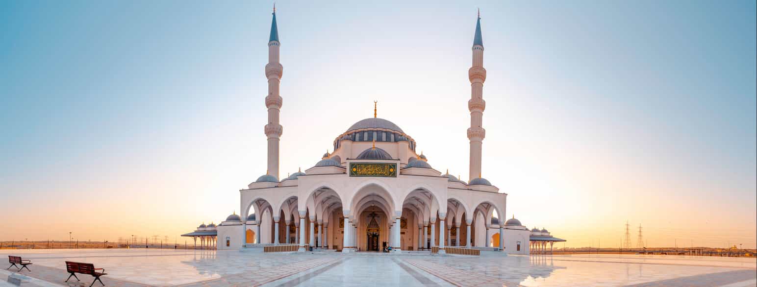 Fotografi av en stor, hvit moské i et helt flatt landskap. Moskeen har to høye, spisse tårn, og på midten har den en blå kuppel. Plassen foran moskeen er dekket av glatte fliser.