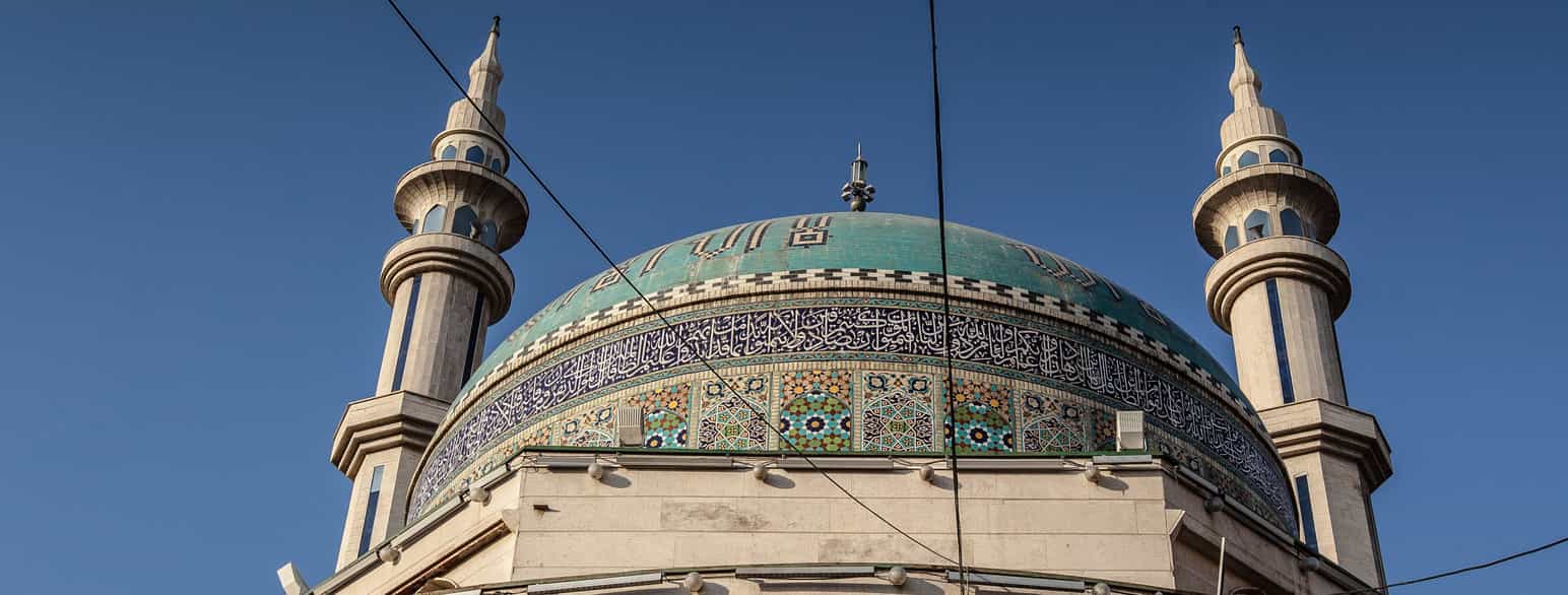 Fotografi av toppen av en moske. Man ser kuppelen, som er dekorert med mosaikk, og de to spisse tårnene, ett på hver side.