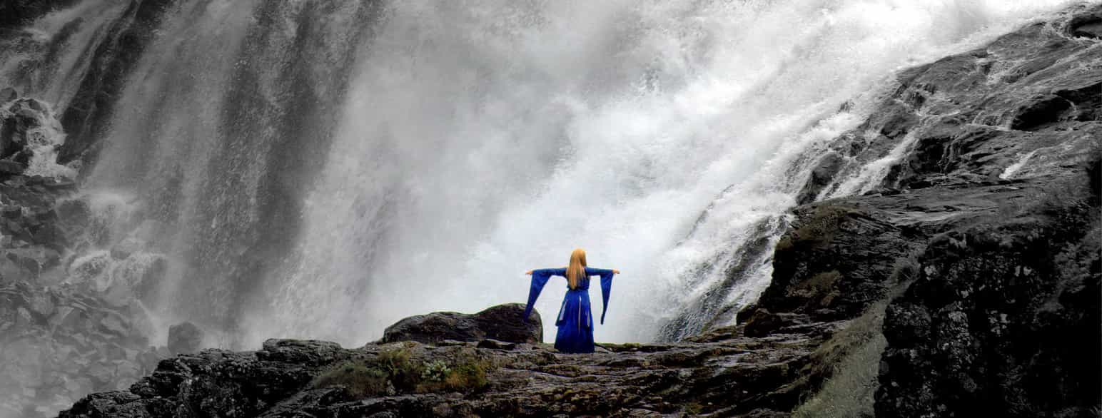 En kvinne i blå kjole, med lange stoffbiter fra ermene, står med ryggen til ved en stor, vill foss. Man kan såvidt se at hun har hale. Foto