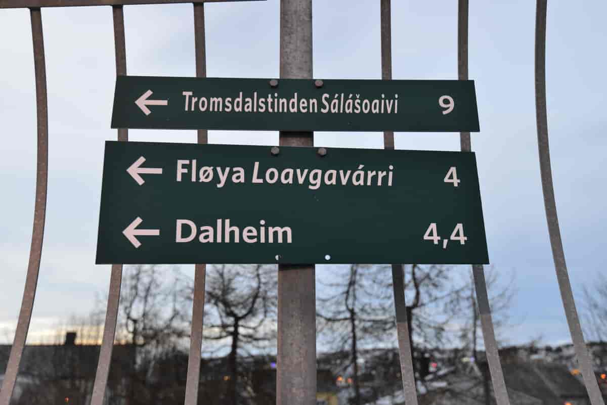 Foto av to skilt med stedsnavn på, festet på en stolpe. På det øverste står det Tromsdalstinden Sálášoaivi 9. På det nederste står det Fløya Loavgavárri 4, og under der står det Dalheim 4,4.