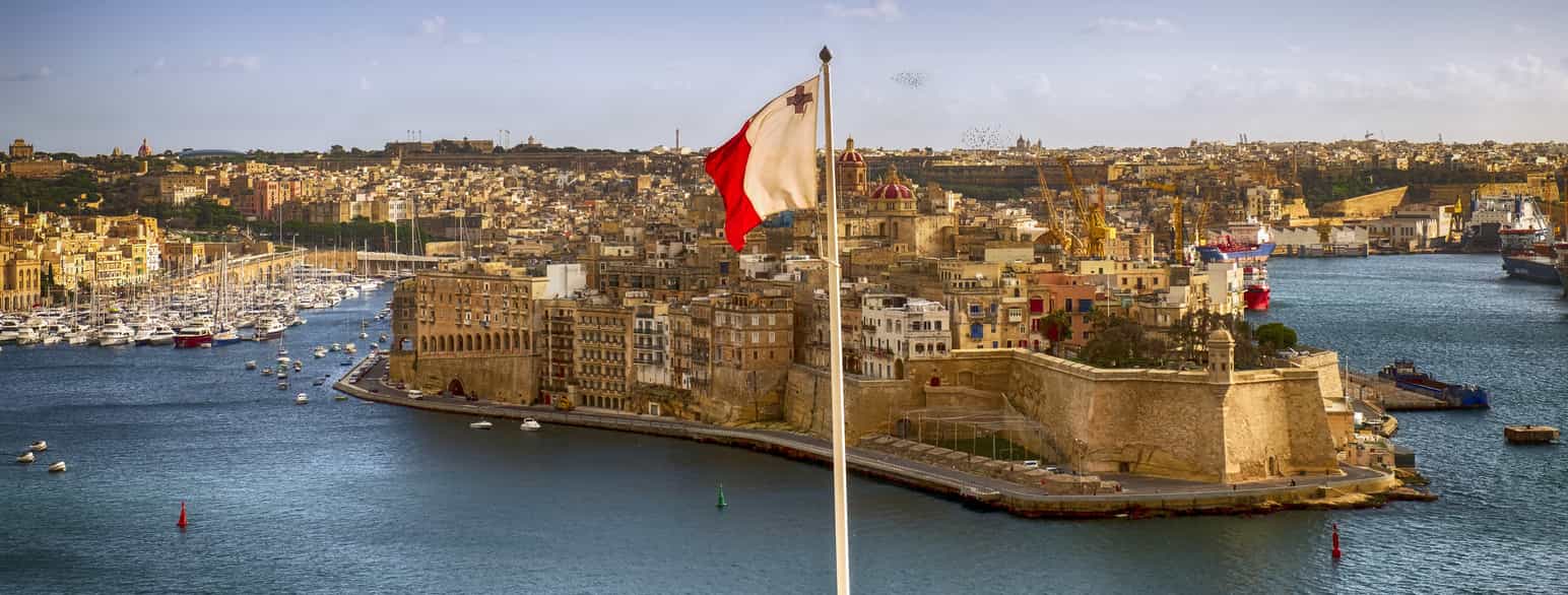 Utsikt over en by sett fra sjøen. I forgrunnen en flaggstang med et maltesisk flagg som vaier i vinden. Midt i bildet er en odde med en gammel festning ytterst. Festningen har høye murer i lys stein. Innenfor festningen er det mer moderne høyhus. Foto