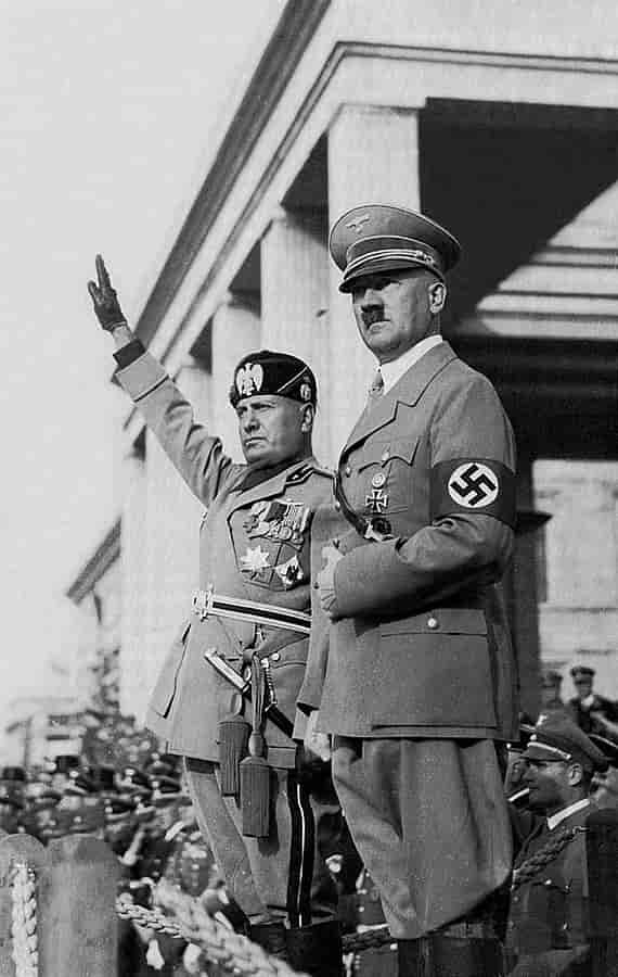 Svart-hvitt foto av Mussolini og Hitler i ulike uniformer. Mussolini (til venstre) holder høyre arm og hånd rett opp i lufta som en hilsen. De to mennene står oppe på en plattform, nedenfor i bakgrunnen står flere menn i uniformer.