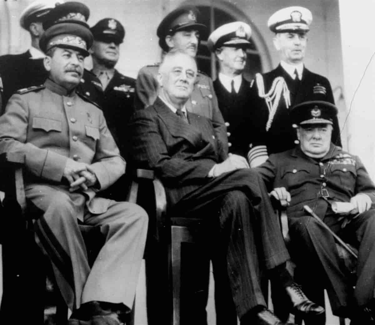 Svart-hvitt-foto av de allierte. Stalin har uniform og en stor bart. Roosevelt har dress, blanke sko og runde briller. Churchill har uniform og en stokk i fanget. De sitter på stoler, bak dem står flere andre menn i forskjellige uniformer.