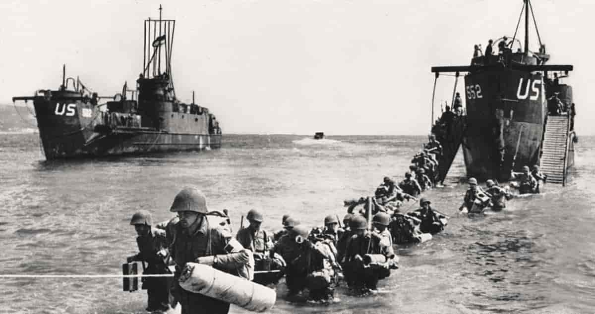 Svart-hvitt foto av to krigsskip som ligger på vannet. Nedenfor og på vei vekk fra det ene skipet er en lang rekke soldater i uniform med hjelmer og oppakning. De har vann omtrent til livet.