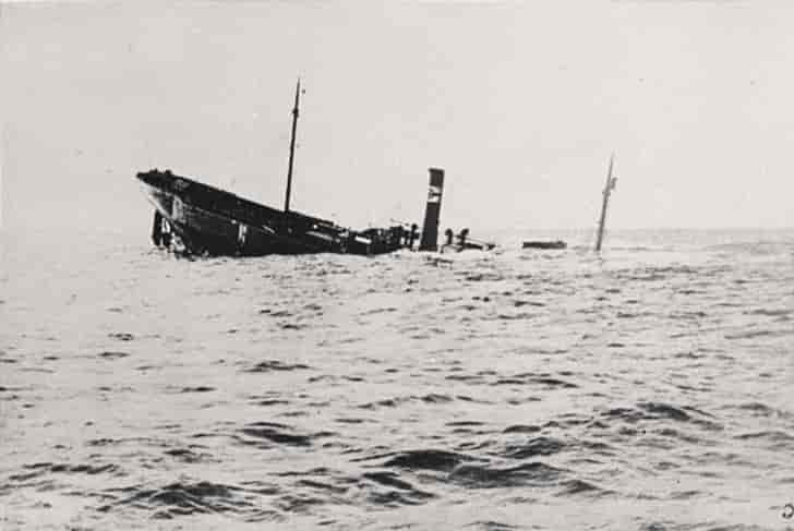 Svart-hvitt foto av et skip med skorstein og to master på vei ned i havet. Bare den ene enden, skorsteinen og de to mastene stikker opp. Resten av båten er under vann.