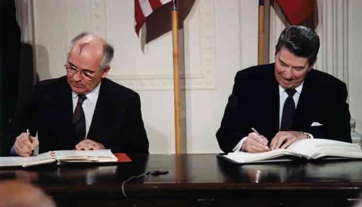 Gorbatsjov og Reagan sitter bak et skrivebord med hver sin åpne perm foran seg, og hver sin penn i hånden. Foto