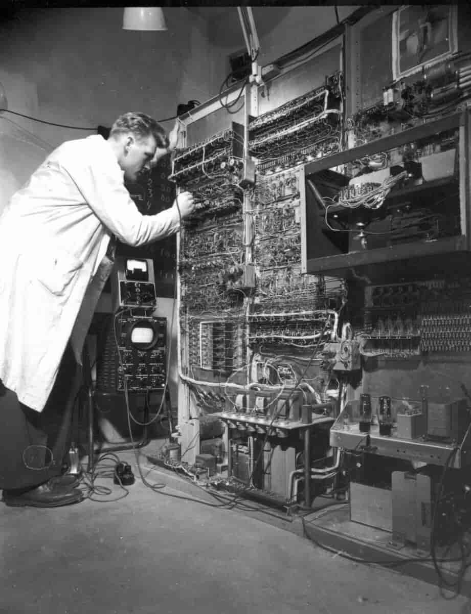 en mann i hvit frakk står foran en stor datamaskin med mange knotter og ledninger.