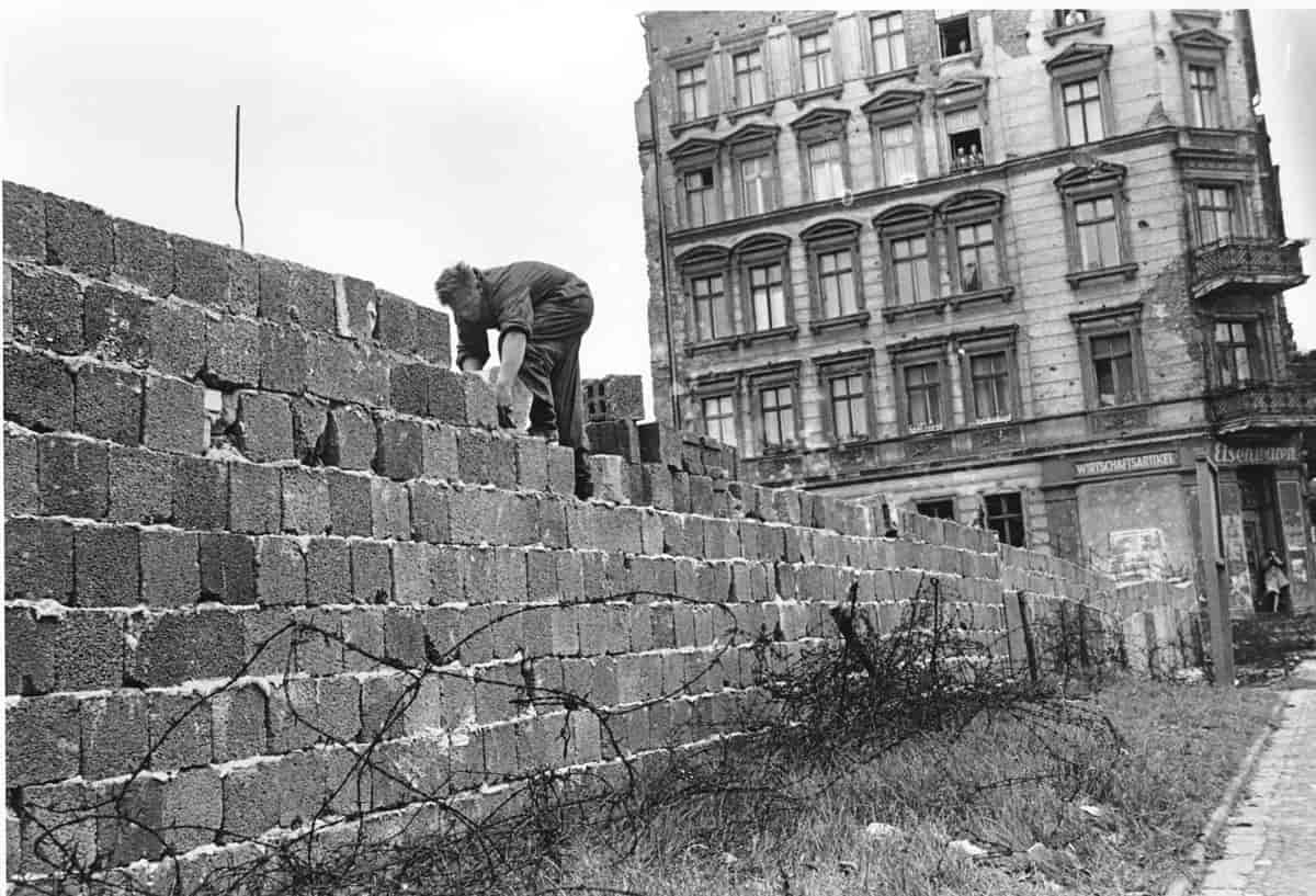 En mann bygger en mur ved siden av en bygård. På bakken ligger piggtråd. Foto