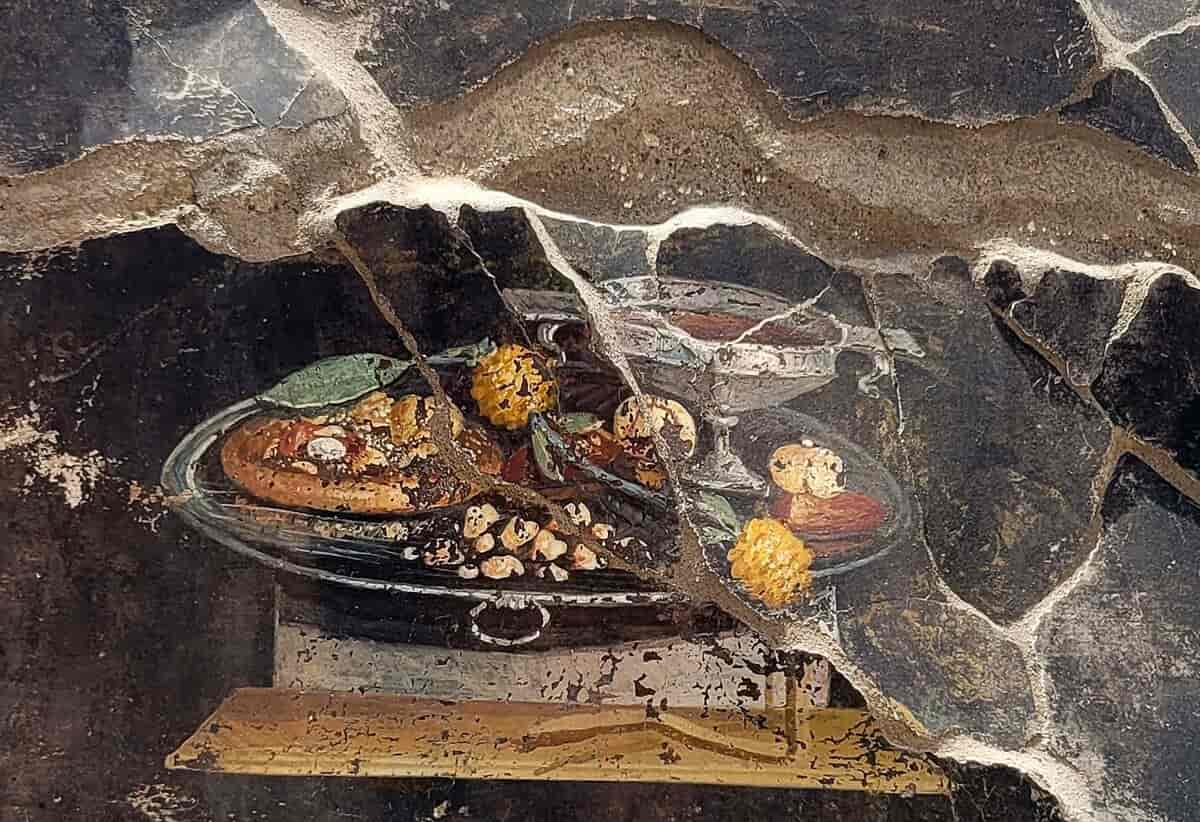 Et veggmaleri av et stort rundt fat med forskjellige matvarer på. Til venstre på fatet ligger det som ser ut som en liten rundt pizza. Foto fra Pompeii