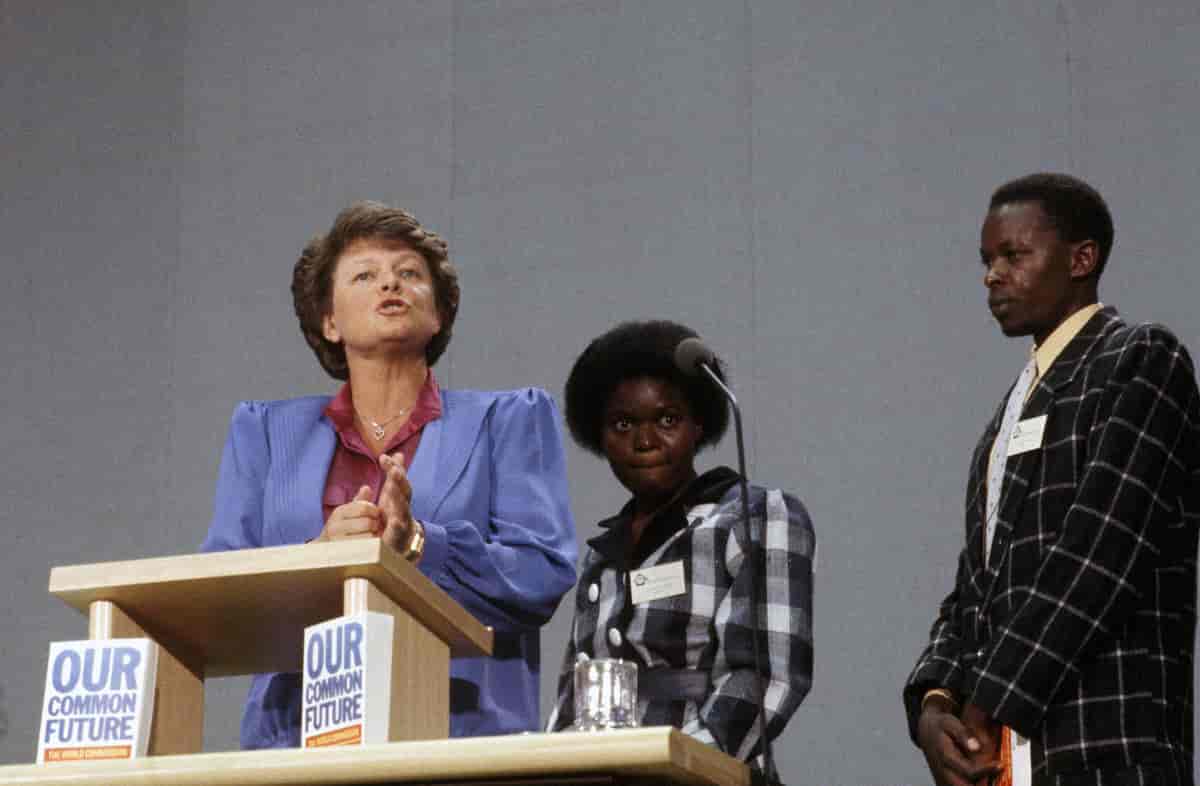 Brundtland på talerstolen i blå dressjakke og rød skjorte. Til høyre for henne to ukjente personer. 
