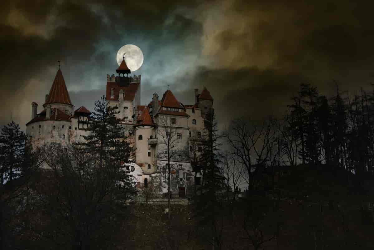 Fullmåne over et gammelt slott med tårn og spir. Det er mørkt og noen lave skyer henger delvis foran slottet og månen. 