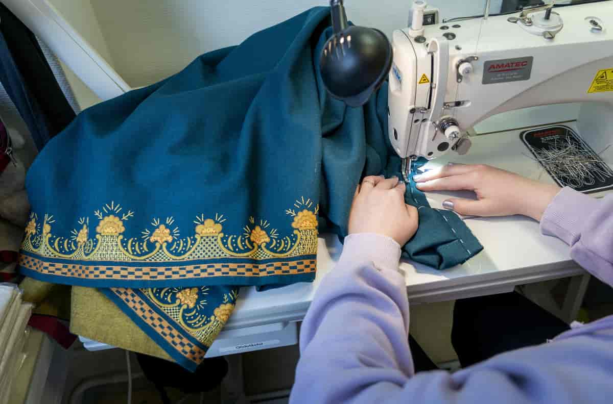 Armene til en person som sitter foran en symaskin. Hun holder på å sy i et blått bunadskjørt med broderier nederst. Foto