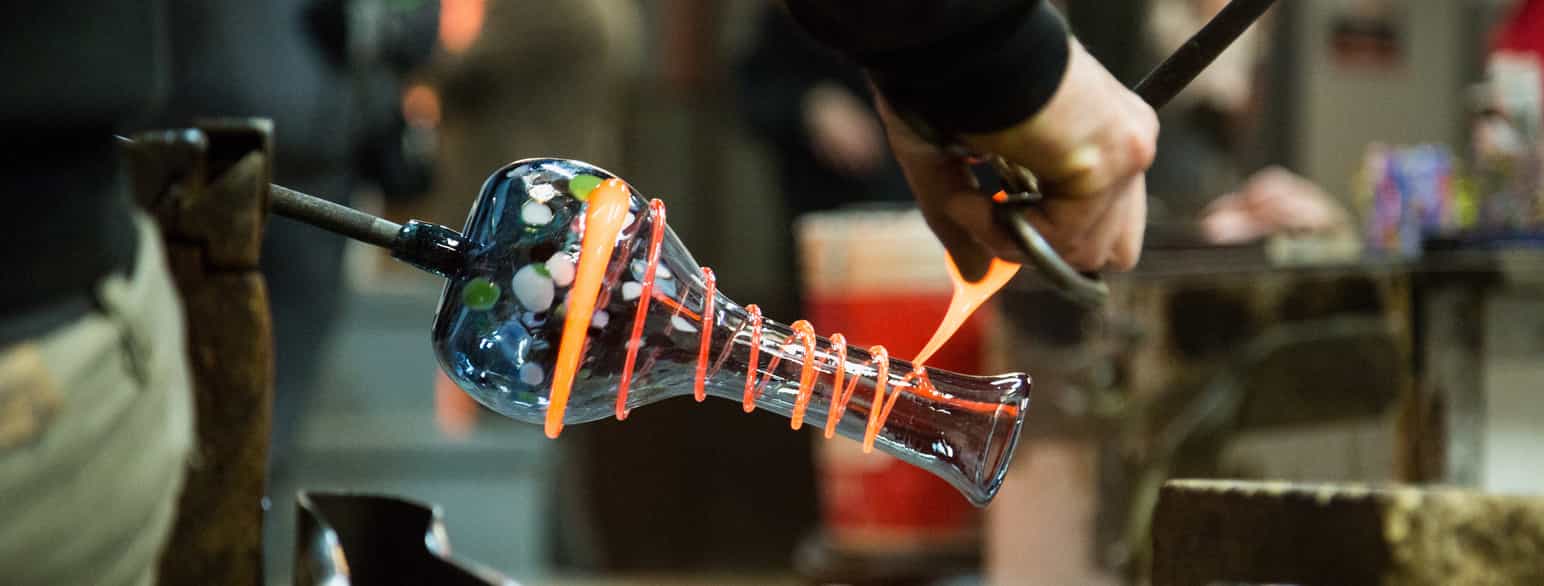 En mann bruker jernverktøy for å spinne en tråd av flytende farget glass rundt en vase av klart glass. Foto