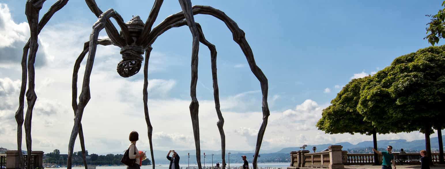 Foto av ein stor edderkoppskulptur i ein by. Ei kvinne går forbi, ein mann tek bilde avskulpturen med mobilen.