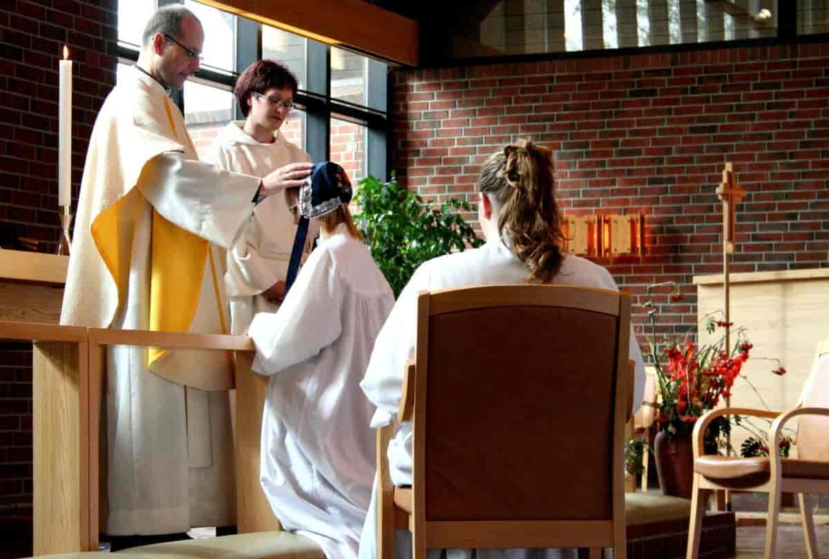 En kvinnelig og en mannlig prest står foran to konfirmanter i en kirke. Den ene konfirmanten kneler foran en av prestene, som har hånden på hodet hennes. Den andre konformanten sitter på en stol. Begge konfirmantene har på seg hvite kjortler.