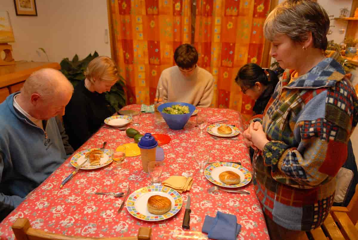 Et bord er dekket på til fem personer. Det er tallerkener med mat, men ingen har begynt å spise. Fire personer sitter rundt bordet med hodene bøyd. En kvinne har reist seg og står med hendende foldet og hodet bøyd. 