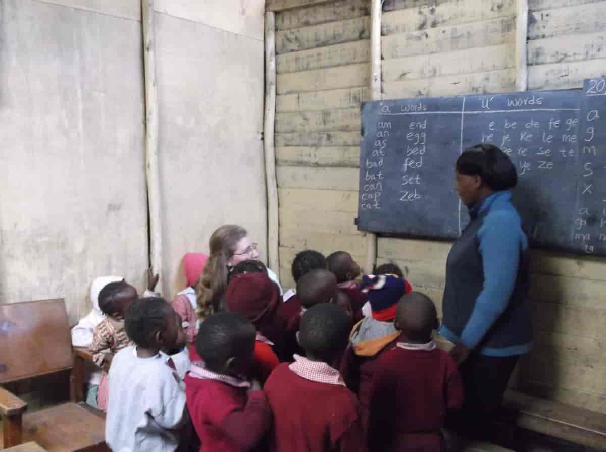 Små skoleelever står og ser på læreren sin. Hun står foran en tavle hvor det er skrevet engelske ord. Midt blant barna sitter det en kvinne med lyst hår og briller.