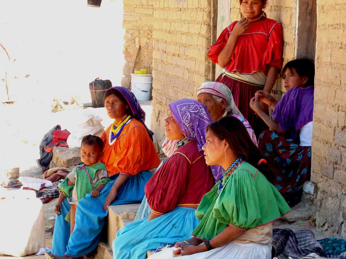 En gruppe kvinner i fargerike klær sitter i skyggen utenfor et hus. En av dem har et barn i fanget.