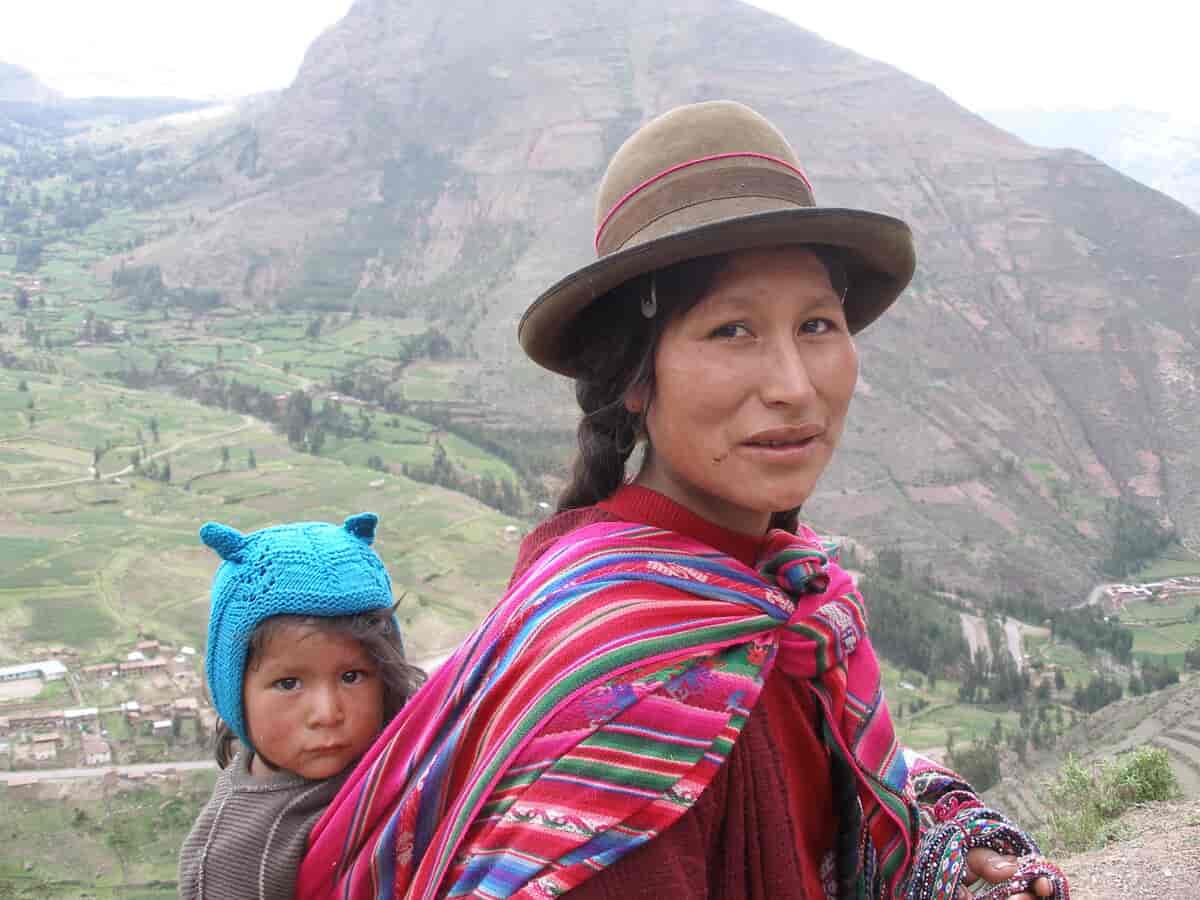 Foto av en kvinne som bærer et barn på ryggen og har knyttet et fargerikt tøystykke rundt skuldrene.  Hun står i et fjell-landskap.