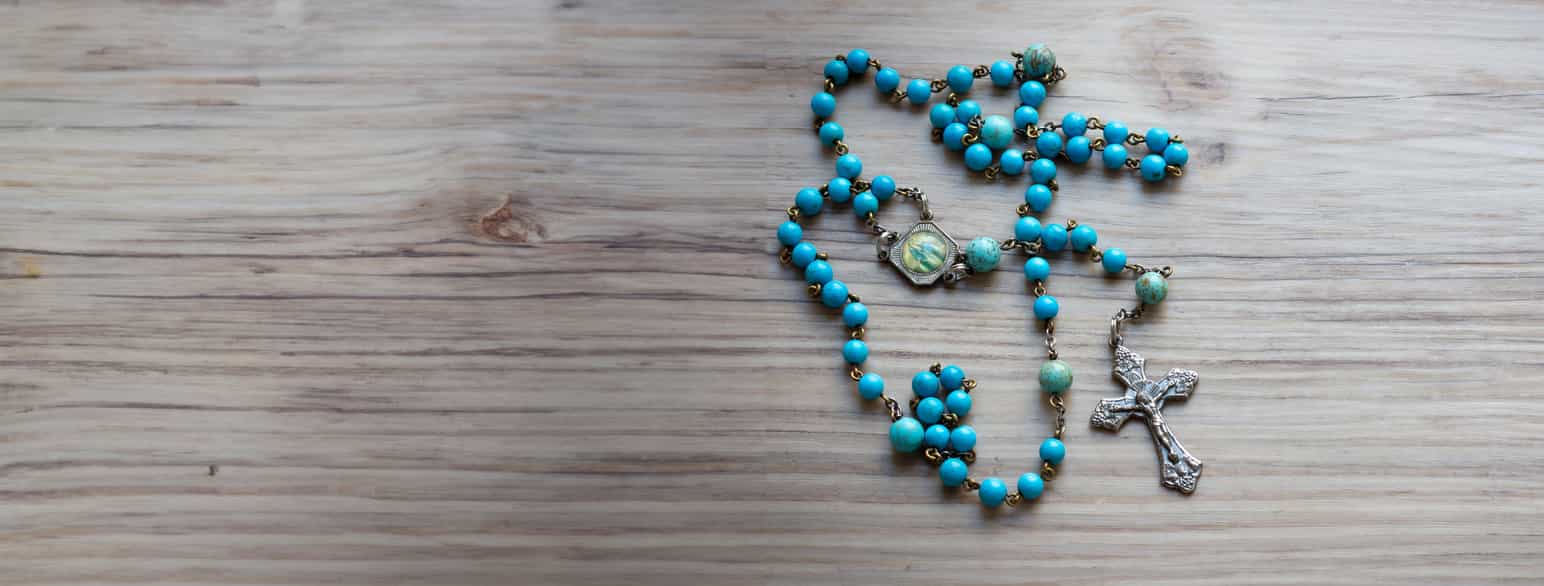 Foto av et blått bønnekjede som ligger på et bord. I den ene enden er det et kors. Man kan også se et en liten plate med et bilde av Maria, moren til Jesus, som er festet på kjedet.