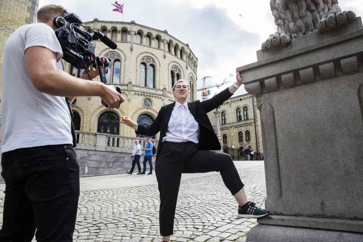 Maria Stavang i kostyme står med det ene beinet opp på en av løvestatuene foran Stortinget. En mann med filmkamera filmer henne.