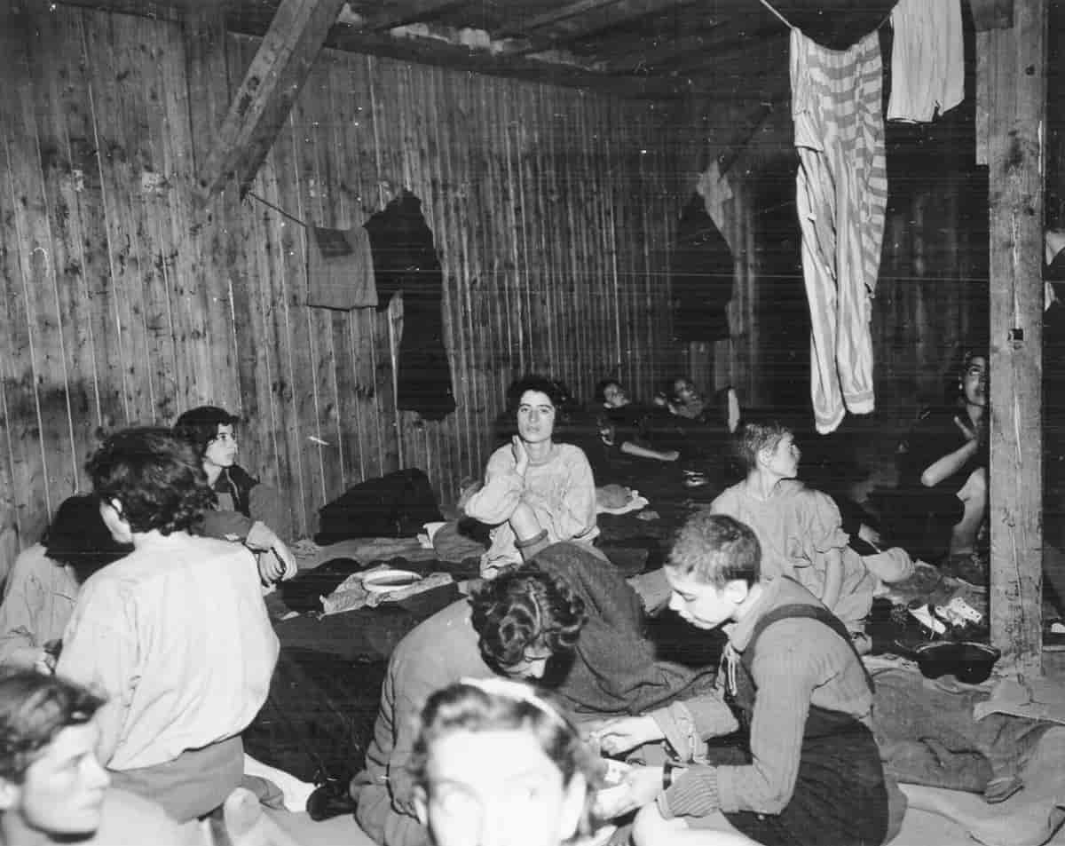 Sort-hvitt fotografi av kvinner som sitter på gulvet i et lite rom. De har tepper rundt seg. Fra taket henger klær til tørk.