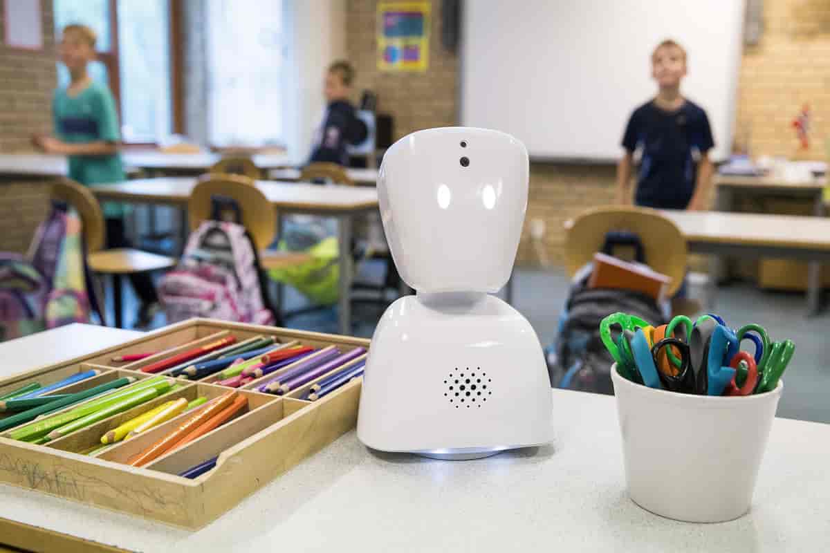 En liten hvit robot står på en pult i et klasserom. Bak roboten står det noen barn.