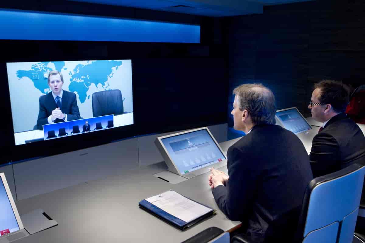 Bildet viser to menn som sitter og ser på en videoskjerm på veggen foran dem. På skjermen ser man et stort bilde av en mann som snakker og et mindre bilde av en mann som lytter. Foran de to mennene er det også små skjermer som viser deltakere på konferansen.