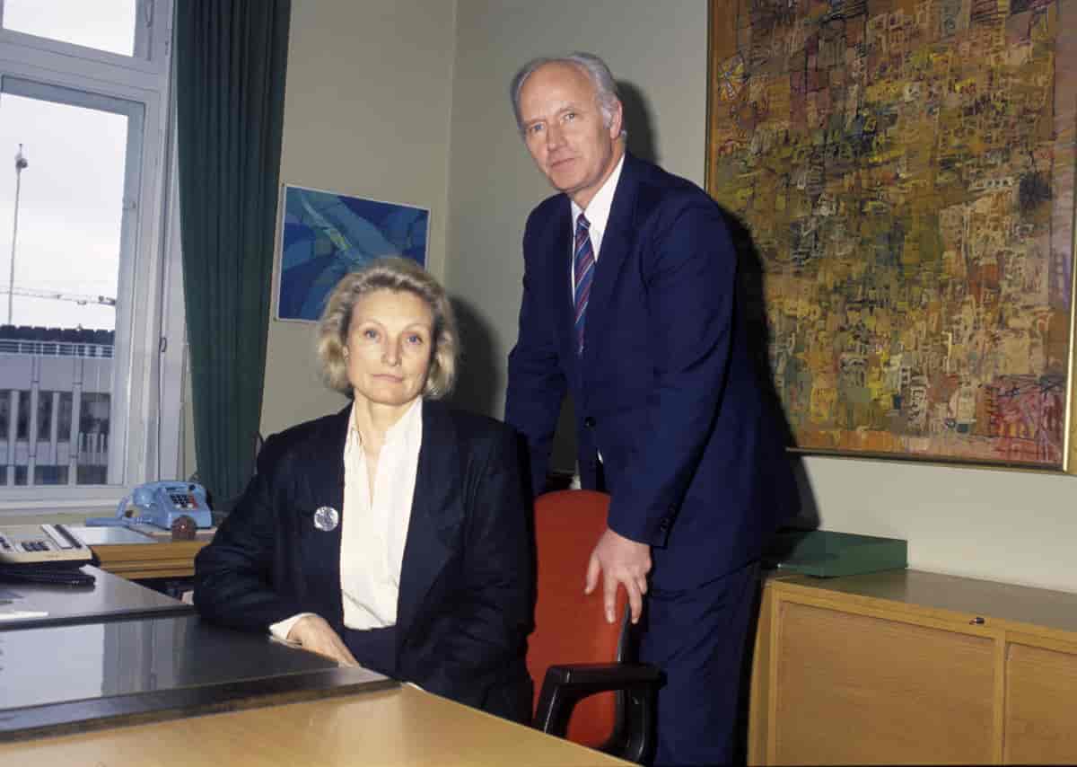 Helga Hernes sitter ved en pult, Stoltenberg holder i stolen hennes. Begge ser i kamera. 