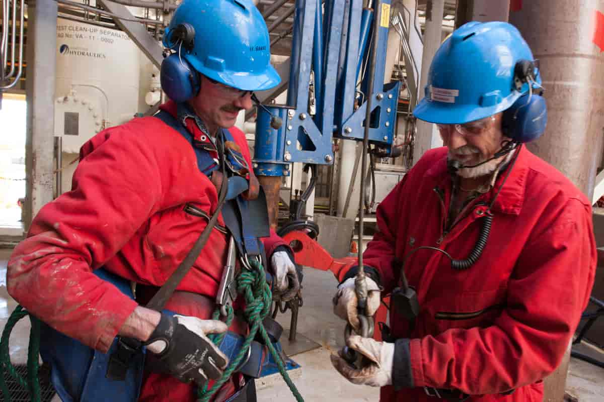 Foto av to menn med arbeidsklær og hjelmer på en oljeplattform. 