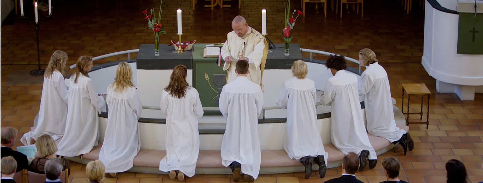Flere hvitkledte ungdommer sitter på kne ved alterringen. En prest står i midten. Han har hånden på hodet til en av ungdommene.