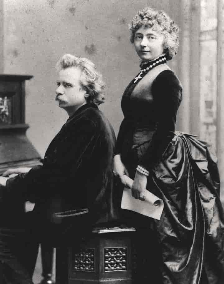 Svart/hvitt bilde av Edvard Grieg ved pianoet. Ved siden av ham står kona Nina.