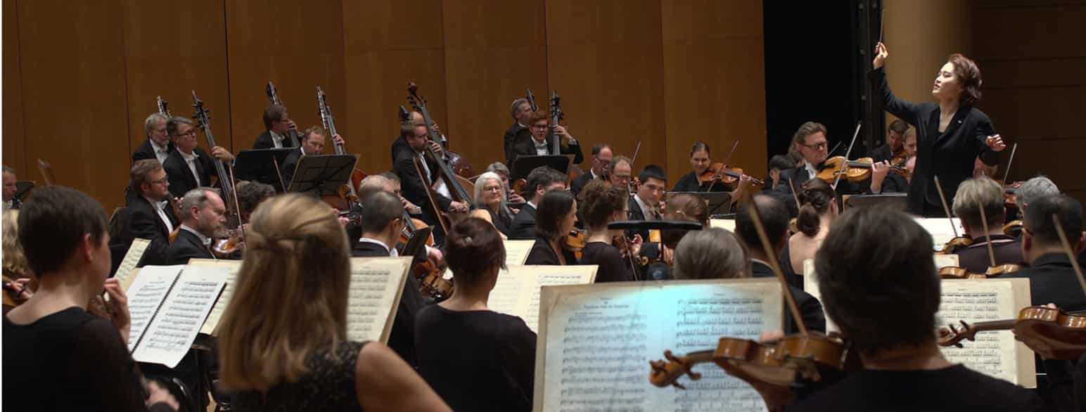 Et orkester sett bakfra med en kvinnelig dirigent i bakgrunnen.