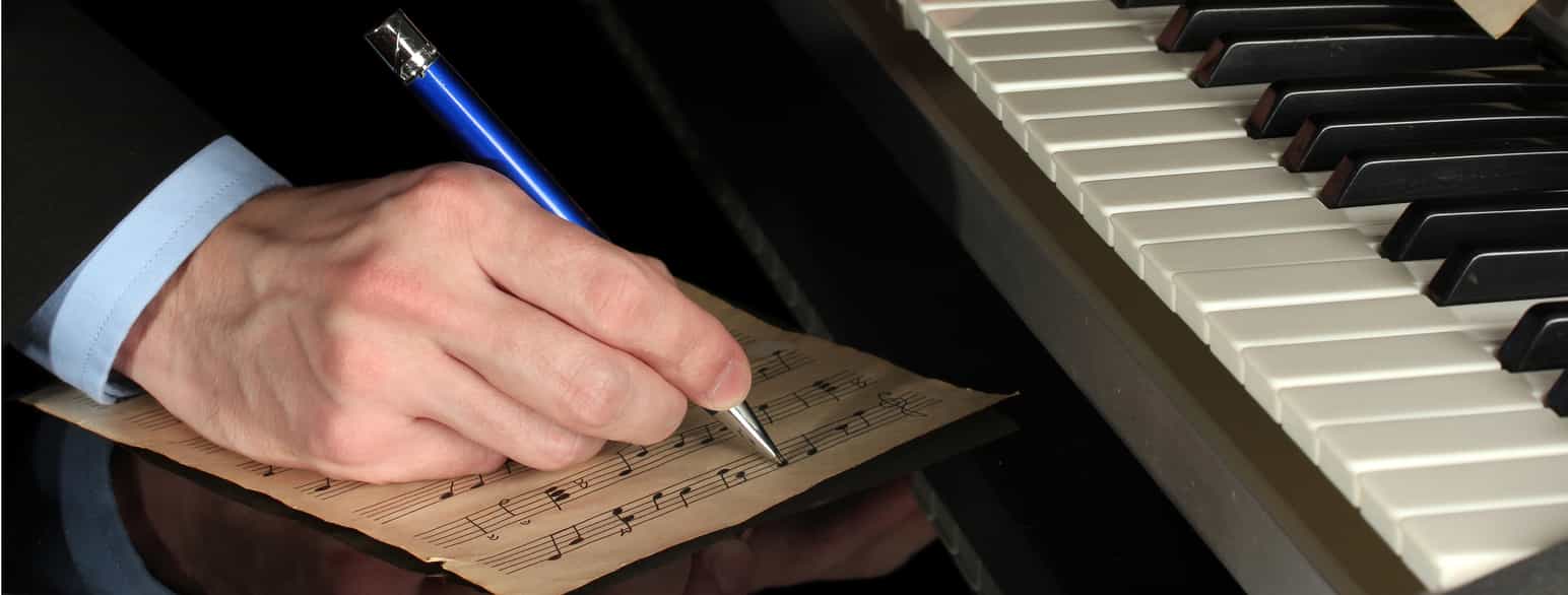 Foto av en hånd som skriver noter, og i bakgrunnen svarte og hvite tangenter på et piano. 
