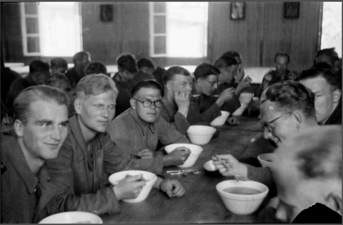 Sort-hvitt fotografi av en spisesal der mange unge menn sitter ved et langbord og spiser suppe.