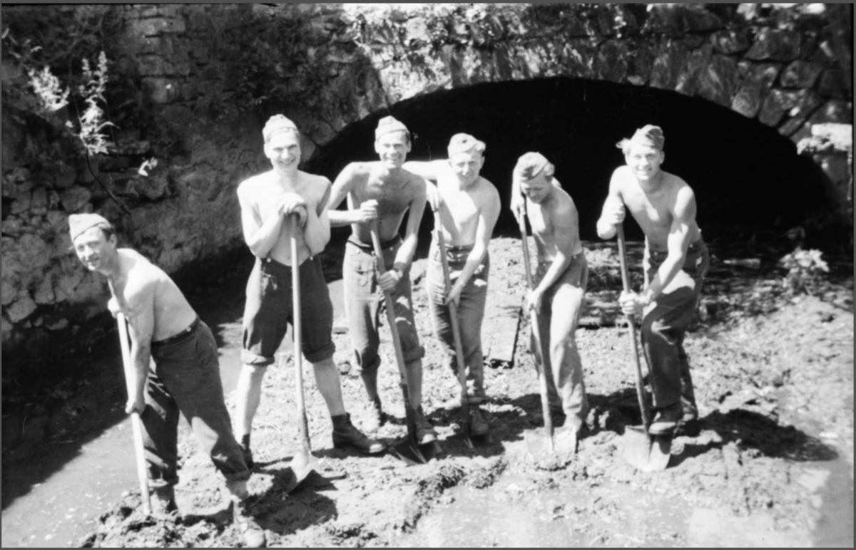 Sort-hvitt fotografi av seks unge menn med hver sin spade. De står ved en gjørmete elvebredd og ser på fotografen, flere av dem smiler.