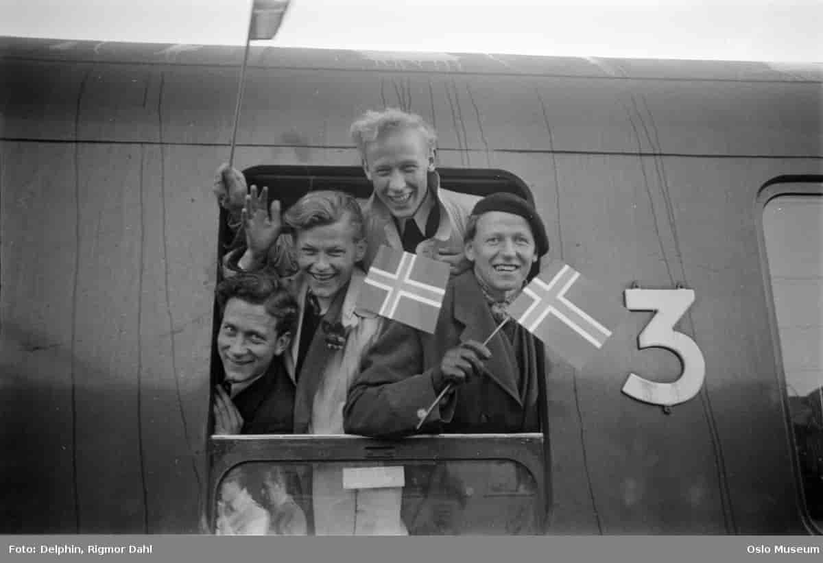 Sort-hvitt fotografi av en togvogn med fire unge menn som stikker hodene ut av togvognas åpne vindu. De smiler og vinker, og to av dem har norske flagg i hendene.