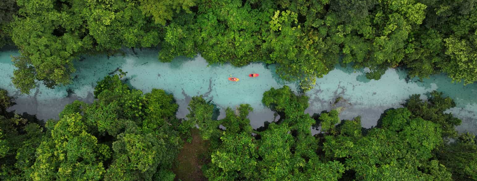 Dronefoto av elv med tett grønn skog på hver side. På elven er det to bittesmå båter med en person i hver.