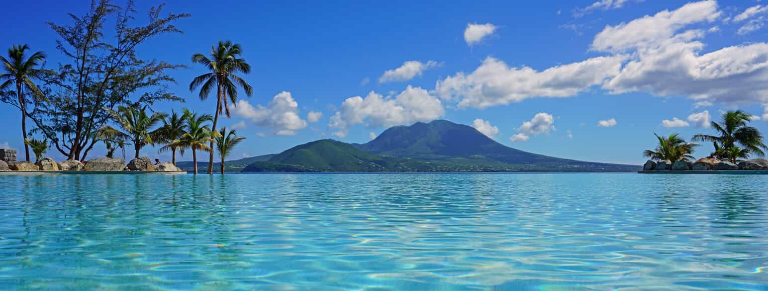 I forgrunnen palmer og blått hav, i bakgrunnen en vulkan på en annen øy.