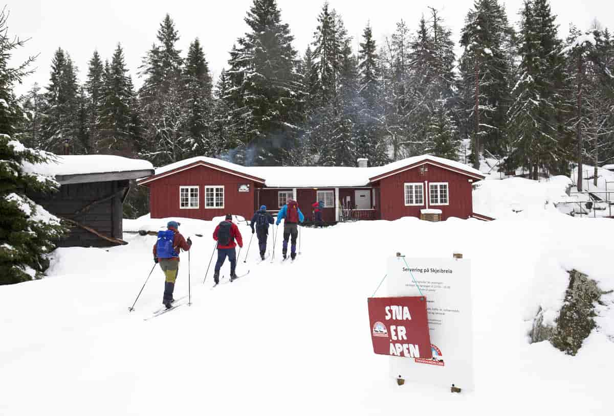 Fem skigåere på vei mot en stor rød hytte. Skilt i forgrunnen der det står: "Stua er åpen". 