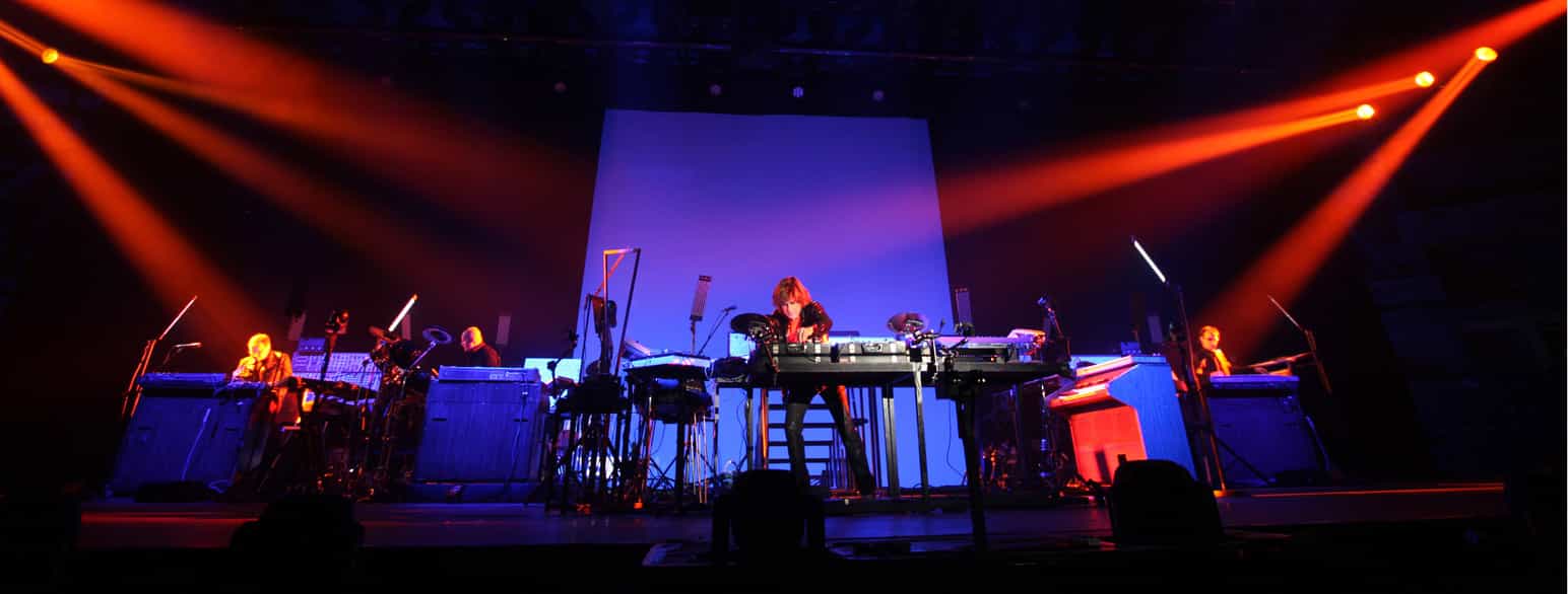 Musikeren på en stor scene med mange datamaskiner og elektronisk utstyr i forskjellige størrelser foran seg.
