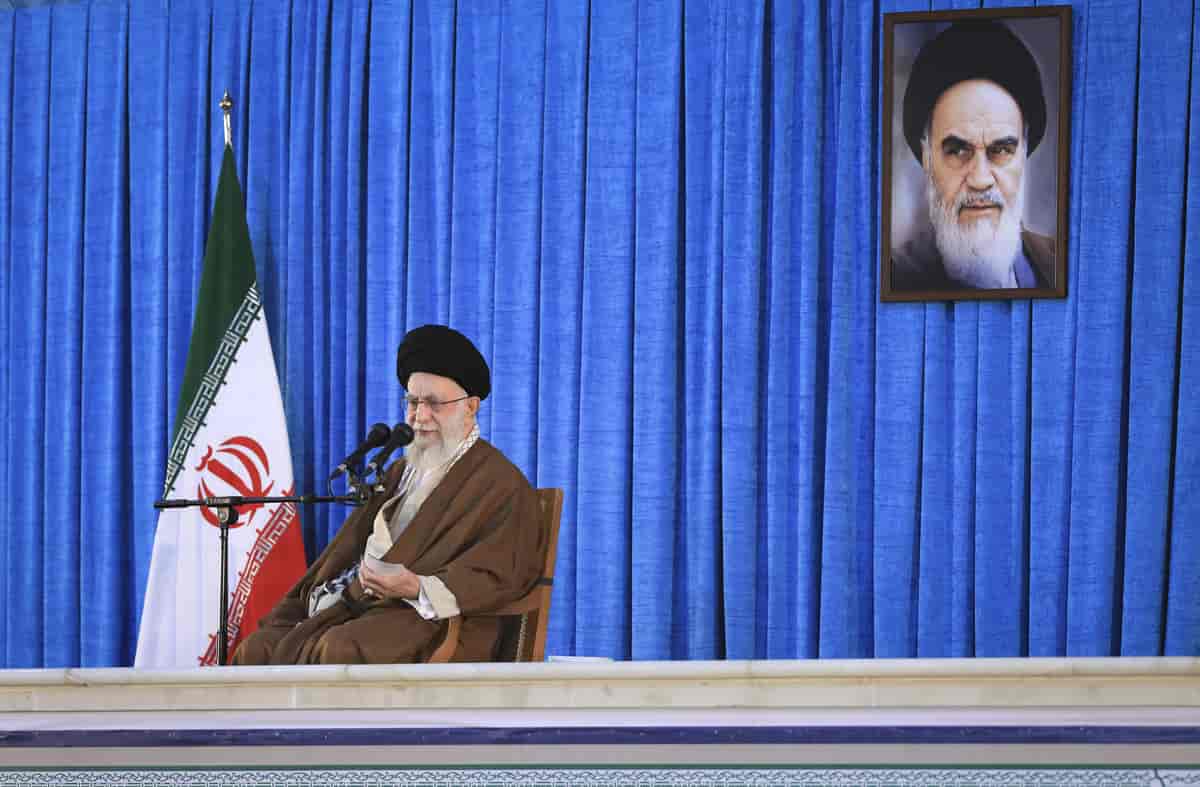 Ali Khamenei, kledd i turban holder en tale. Et bilde av Ruhollah Khomeini henger på veggen bak ham.