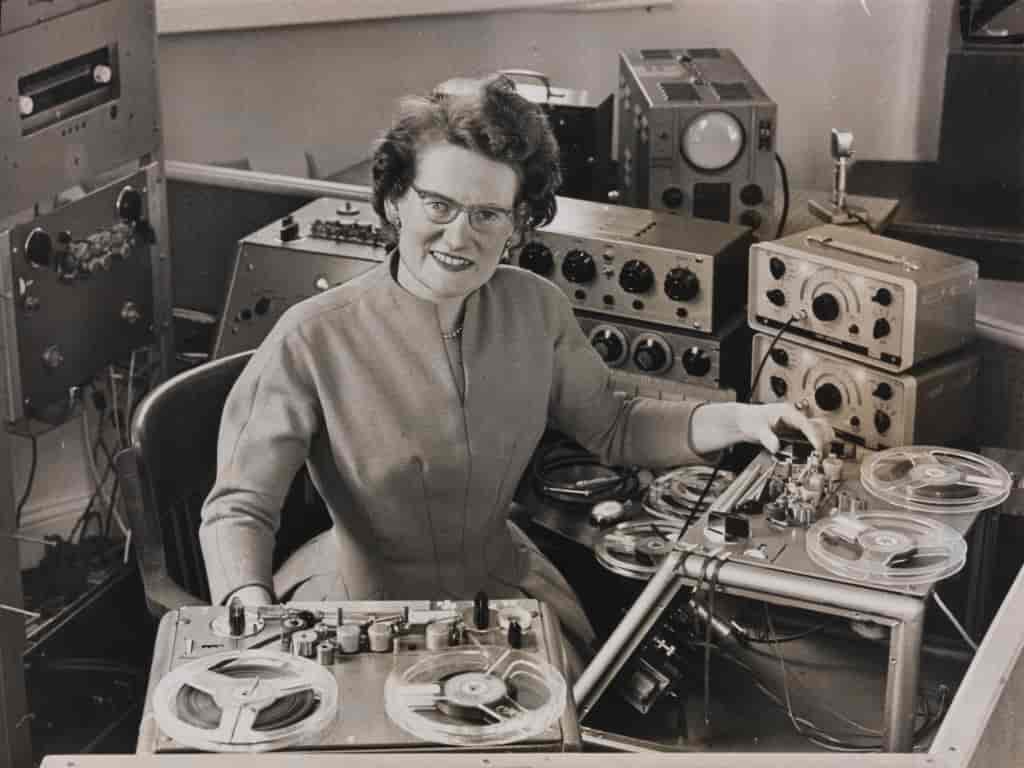 Svart/hvitt bilde av en kvinne som sitter omgitt av maskiner som kan lage elektronisk lyd, for eksempel en båndopptager.