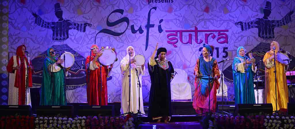Flere kvinner i fargerike kjoler synger og spiller på en scene. På veggen bak står det forskjellig tekst, som sufi. Det er fordi gruppen synger og spiller på en sufi-festival.
