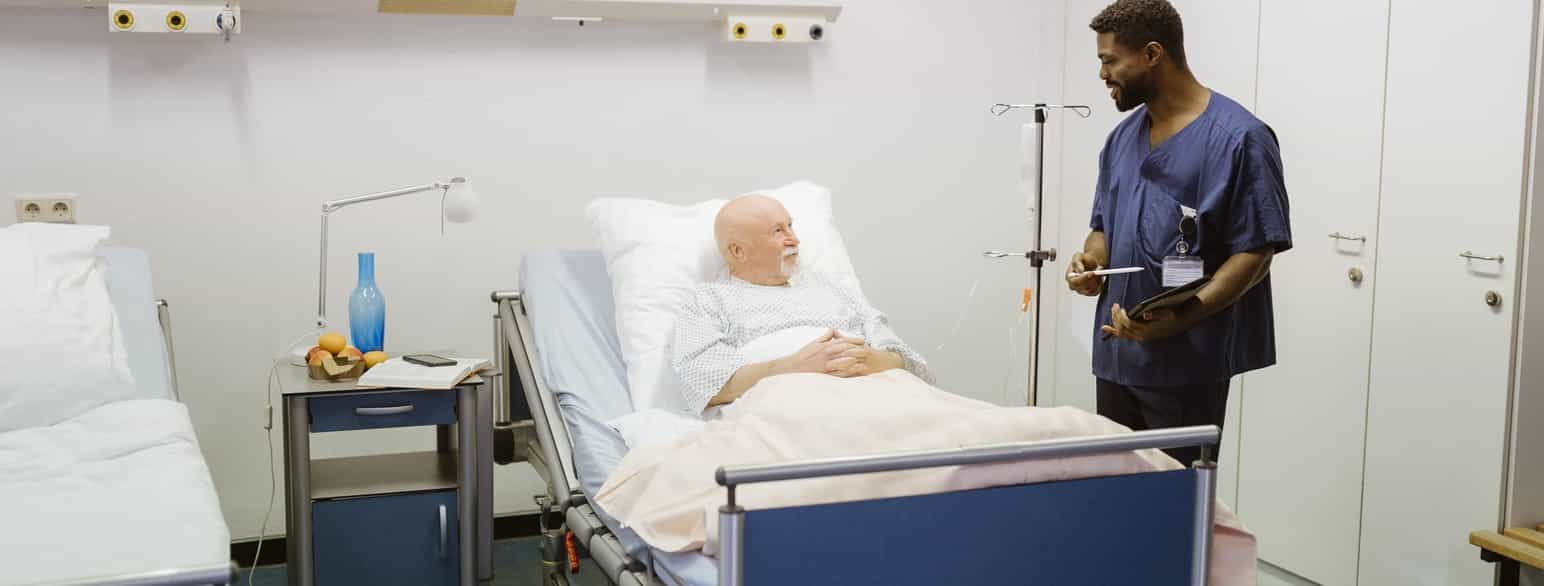 Foto av sykepleier i blå sykehusklær som snakker med en eldre pasient som ligger i en sykehusseng. 
