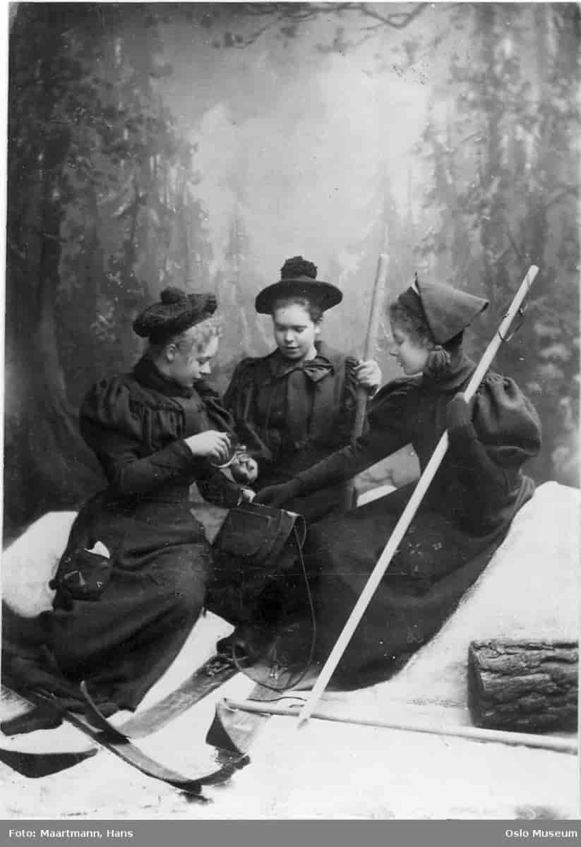 Tre kvinner i pent svart tøy som sitter i snøen. De har treski på beina og en tykk stav hver. Den ene kvinnen holder en lommelerke og tømmer i et lite glass. 