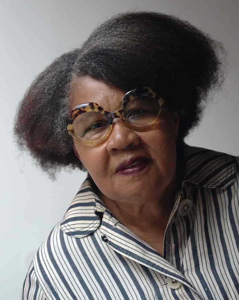 Halvnært foto av Jamaica Kinkaid. Hun bærer store markante briller med tykk innfatning. Håret er naturlig gråsort, sveisen er kink afrobob. Bomullskjorten er hvit med blå striper og avrundede snipper.