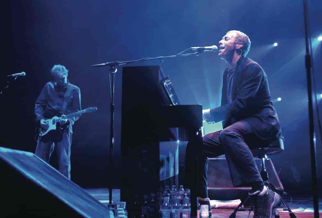 En musiker spiller piano og synger inn i en mikrofon. Til venstre for ham er det en gitarist.